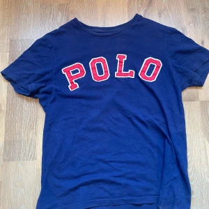 Blå t-shirt från Polo Ralph Lauren - Mörkblå t-shirt från Polo Ralph Lauren med stora röda bokstäver 'POLO' framtill. Tröjan är storlek S och passar XS/S Hör av dig vid några funderingar!