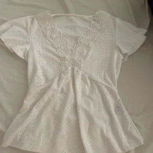 Vit blus med spets och v-ringning - Supersöt vit blus med korta ärmar, djup v-ringning och detaljerad spets längs urringningen. Blusen har broderade hålmönster över hela tyget och en lätt utsvängd passform. Perfekt för dig som gillar romantiska och feminina plagg.