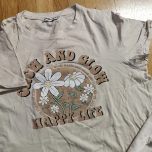 Beige t-shirt med blommor från FB New Yorker - Beige t-shirt från NewYorker med coolt tryck av glada blommor och texten 'Grow and Glow Happy Life' i retrostil. Skön passform och rund halsringning, perfekt för en chill och trendig look. Hör av er om ni vill ha fler bilder eller har frågor💕💕💕