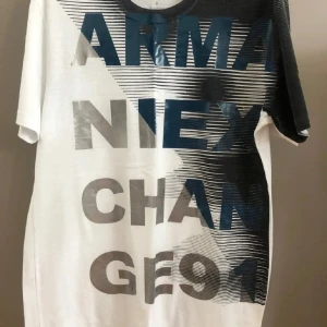 t-shirt Armani Exchange - Snygg t-shirt från Armani Exchange i vitt och blå med stora tryckta bokstäver och grafiskt mönster framtill. Klassisk rund hals och korta ärmar. Tillverkad i mjuk bomull, perfekt för en trendig och avslappnad look.