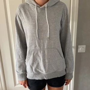 Grå hoodie från H&M med luva  - Enkel och stilren grå hoodie från H&M i storlek S. Tröjan har huva med vita snören, känguruficka framtill och långa ärmar. Perfekt för chill dagar eller när du vill ha en avslappnad look. Tillverkad i mjuk bomullsblandning för extra komfort. Säljer pga inte kommit till användning.