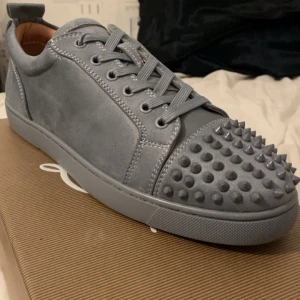 Gråa Louboutin sneakers med nitar - Snygga grås skor från Louboutin med ikonisk röd sula och nitar på tåpartiet. Tillverkade i Italien och tillkommer med kvitto bevis samt dustbag och Christian Louboutin kort. Nypris: 9500, om du har funderingar är du välkommen att fråga! Bilderna tagna på exakt samma sko fast i storlek 44, skorna för salu är storlek 43.
