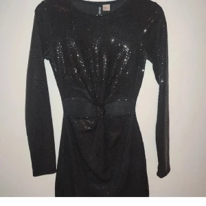 Svart glittrig klänning med knyt - Svart kortklänning från hm med glittrigt tyg och lång ärm. Klänningen har ett snyggt knyt i midjan som ger en cool siluett. Perfekt för dig som vill sticka ut med lite extra glitter.