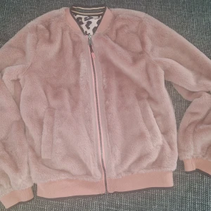 Rosa teddyjacka & leopard bomberjacka - Tvåsidig jacka från ebbe kids! Ena sidan är en fluffig rosa teddyjacka med broderad text 'FEELING FINE' på ryggen, andra sidan är en bomberjacka med leopardmönster i rosa och brunt. Båda sidor har dragkedja och ribbade muddar. Perfekt för dig som vill variera din stil.