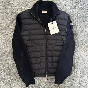Navy Moncler pufferjacka M - Snygg svart pufferjacka från Moncler i storlek M. Jackan har quiltad framsida i syntetmaterial och stickade ärmar och rygg. Hög krage, dragkedja framtill och diskret Moncler-logga på ärmen. Perfekt för dig som vill ha en stilren och sportig look.
