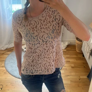 Ljusrosa spetsblus med knappar bak - Säljer en ljusrosa blus i spets med korta ärmar och rund hals. Blusen har fina knappar längs ryggen och ett genombrutet spetsmönster som ger en extra touch.