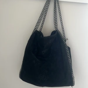 Svart axelväska med kedjedetaljer - Säljer en svart axelväska med coola silverfärgade kedjor som axelremmar och längs kanterna. Väskan är i glansig mocka-look och har dragkedja upptill. Storlek som en tygpåse. 