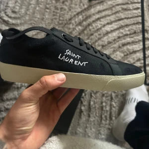 Svarta Saint Laurent sneakers strl 41 - Svarta sneakers från Saint Laurent med vit sula och handskriven logga på sidan. Skorna har snörning, rund tå och är tillverkade i canvas och skinn. Klassisk och stilren modell med diskreta detaljer och guldtext på plösen. Perfekta för dig som gillar lyxig streetstyle.