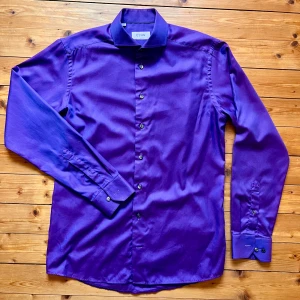 Lila skjorta från Eton i bomull - Snygg lila skjorta från Eton i slim fit med klassisk krage och svarta knappar. Skjortan är tillverkad i bomull och har långa ärmar med manschett. Perfekt för dig som vill sticka ut med färg och stil.