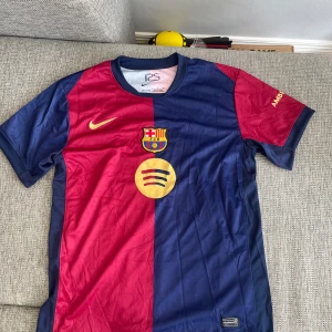 FC Barcelona matchtröja Nike blå/röd - Säljer en officiell Barcelona matchtröja från Nike med klassisk halva-halva design i blått och rött. Tröjan har klubbmärke, Nike-logga och Spotify-sponsor framtill samt UNHCR-tryck bak. Tillverkad i lätt och ventilerande polyester, storlek L