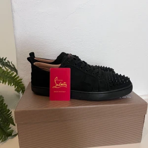 Svarta Christian Louboutin sneakers med nitar - Snygga svarta sneakers från Christian Louboutin i mocka med svarta nitar över tån för en edgy vibe. Skorna har rund tå, snörning och svart platt sula. Insidan är ljusbeige och de har en exklusiv känsla som sticker ut. 900 Kr för dessa skor i 24 timmar sedan återgår priset till 1200 kr