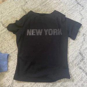 Svart t-shirt med New York-tryck - Svart t-shirt med korta ärmar och texten NEW YORK i glittriga stenar framtill. Snygg och enkel modell som passar perfekt till jeans eller kjol. Materialet känns mjukt och stretchigt, troligtvis bomull eller polyester.