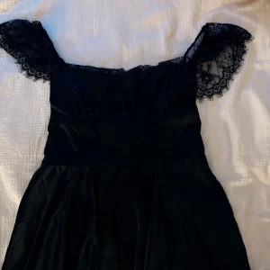 Svart klänning med spetsdetaljer - Supersnygg svart klänning i satinliknande material med spets på axelbanden och omlottkänsla i ryggen. Klänningen har en markerad midja och lätt utställd kjol, perfekt för dig som vill sticka ut med elegans. Knytning i ryggen för extra detalj.