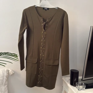 Olivgrön snörad klänning 77FLEA - Olivgrön kortklänning från 77FLEA med långa ärmar och cool snörning framtill. Klänningen har två stora fickor framtill och är gjord i mjukt material med polyester och viskos. Snygg och enkel att styla till sneakers eller boots. Storlek L, upplever den mer som M. Tyg under snörningen så storlek justeras med snörning. Ej använd.