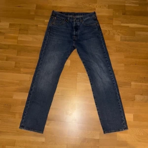 Levi's 501 blå denim - Klassiska Levi's 501 jeans i blå denim med raka ben och normal passform. Byxorna har fem fickor, knappgylf och den ikoniska läderpatchen bak i midjan. Perfekta för dig som gillar tidlös stil och vill ha ett par schyssta jeans i bomull.