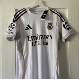 Real Madrid Vini Jr. 7 Adidas tröja - Real Madrid matchtröja med Vini Jr. 7 på ryggen. Vit med svarta detaljer, gula linjer och klubbmärke på bröstet. Adidas-logga, Champions League-märke på ärmen och 'Emirates Fly Better' tryck. Tillverkad i lätt funktionsmaterial (AEROREADY).