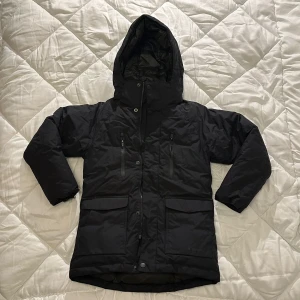 Svart dunjacka från Everest - Säljer en svart dunjacka från Everest med huva och dragkedja. Jackan har flera praktiska fickor och är perfekt för kalla dagar. Den är quiltad för extra värme och komfort. Passar bra för vintersäsongen.