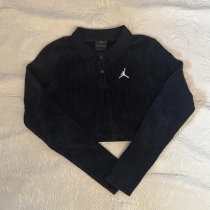 Svart ribbad polotröja från Jordan - Snygg svart croppad polotröja från Jordan med ribbad textur och broderad logga på bröstet. Tröjan har långa ärmar och knappar framtill. Perfekt för en sportig look!