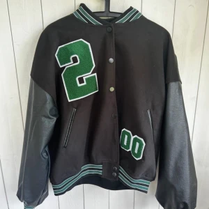 Svart och grön bomberjacka från Boohoo - Säljer en svart bomberjacka från Boohoo med gröna detaljer och stora siffror på framsidan. Jackan har långa ärmar i skinnimitation och stängs med knappar. 