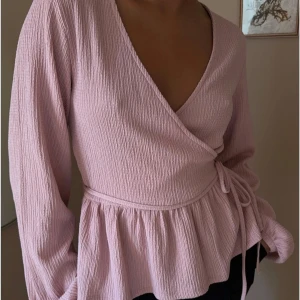Rosa omlottopp - Säljer en söt rosa omlottopp med långa ärmar och knytband i midjan. Toppen har en v-ringad design och är tillverkad i ett mjukt, texturerat material som ger en fin passform.
