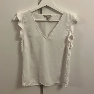 Vit ärmlös topp från H&M - Säljer en stilren vit ärmlös topp från H&M med volangdetaljer vid ärmhålen. Toppen har en v-ringad design och är perfekt för en elegant look. Passar bra till både jeans och kjol.