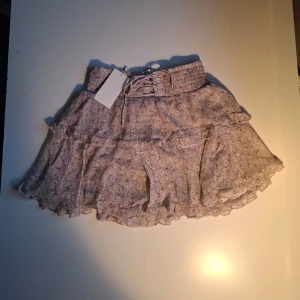 Ljuslila blommig kjol med volang - Charmig ljuslila kortkjol med blommigt mönster och volangdetaljer. Kjolen har en elastisk midja med knytband för justerbar passform. Perfekt för en somrig look! Aldrig använda och lappen kvar!