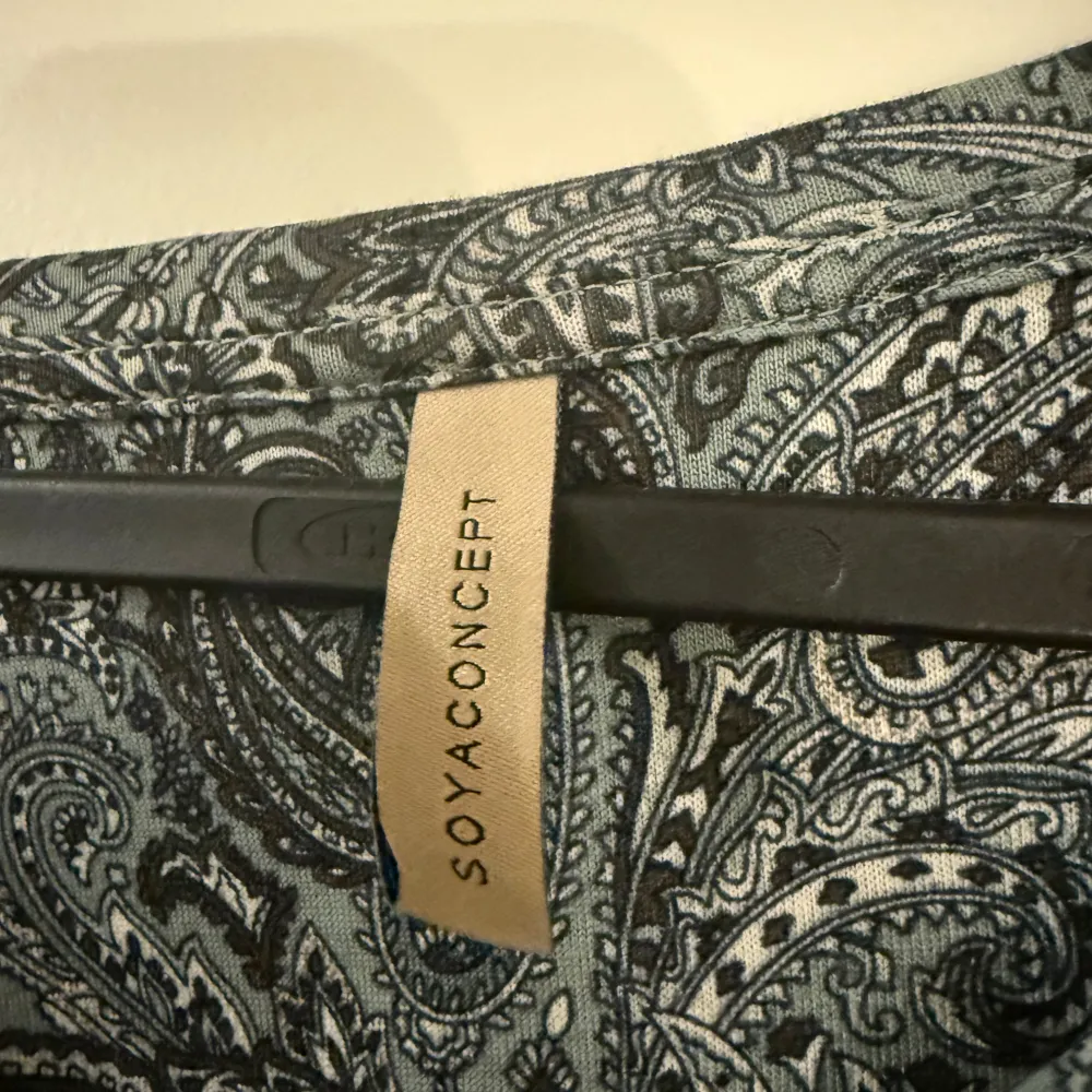 Snygg långärmad topp från Soyaconcept med ett detaljerat paisleymönster i grå och svarta nyanser. Perfekt för en stilren look. Toppen har en v-ringad hals och är tillverkad i ett mjukt material.. Neuletakit & Villapaidat.