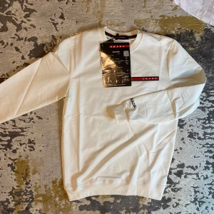 Prada tröja - Säljer en vit tröja från Prada i nytt skick. Tröjan har en klassisk design med logga på bröstet samt ärmen. Perfekt för både vardag och festliga tillfällen. Passar bra till allt, jeans, chinos etc..