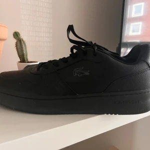 Svarta sneakers från Lacoste  - Snygga svarta sneakers från Lacoste med diskret krokodillogo på sidan. Skorna har en stilren design med snörning och en robust sula. Perfekta för en casual look.