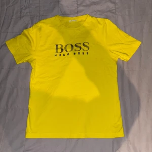 Gul t-shirt från Hugo Boss - Snygg gul t-shirt från Hugo Boss med deras ikoniska logga tryckt i svart på bröstet. Perfekt för en färgglad och stilren look. Kortärmad och i en bekväm passform.