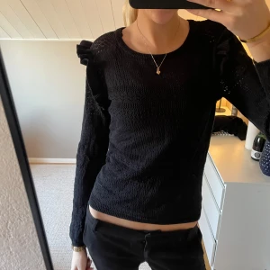 Svart långärmad topp med volangdetaljer - Snygg svart topp med lång ärm och volangdetaljer på axlarna. Toppen har ett fint mönster och är perfekt för en stilren look. Passar bra till både jeans och kjol.
