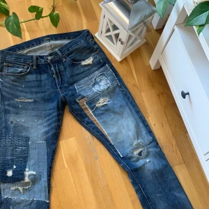 Blå jeans från Polo Ralph Lauren - Säljer ett par blå jeans från Polo Ralph Lauren med coola slitningar och patchwork-detaljer. Jeansen har en klassisk femficksdesign och en vintage look med unika revor och lagningar. Perfekta för en avslappnad stil med en twist.