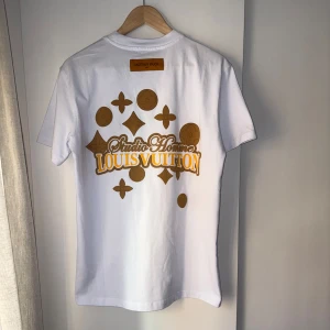 Vit t-shirt från Louis Vuitton - Snygg vit t-shirt från Louis Vuitton med gulddetaljer och logotyp på bröstet. Baksidan har ett stort tryck med Louis Vuitton-mönster i guld. Perfekt för den som vill ha en stilren men ändå iögonfallande look.