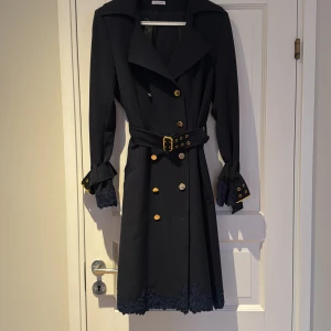 Mörkblå trenchcoat från Ida Sjöstedt - Elegant mörkblå trenchcoat från Ida Sjöstedt med spetsdetaljer och guldknappar. Jackan har ett bälte i midjan och långa ärmar med dekorativa manschetter. Perfekt för en stilfull look. Två knappar har lossnat men en har jag lös och den andra sitter som en extra knapp i kappan. Fint skick!