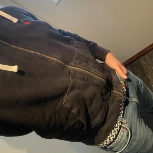 Ralph Lauren - Hoodie - Säljer en svart hoodie från Ralph Lauren med dragkedja och en röd broderad logga på bröstet. Den har en justerbar huva med vita snören och långa ärmar. 