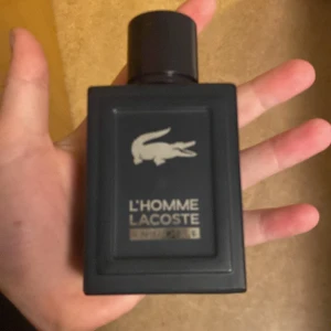 L'Homme Lacoste Parfym - Säljer en stilren och maskulin parfym från Lacoste, L'Homme. Flaskan är svart och rektangulär med en elegant design och det ikoniska Lacoste-loggan. Perfekt för den som vill ha en sofistikerad doft. Ca hälften kvar