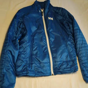 Blå vindjacka från Helly Hansen - Säljer en blå vindjacka från Helly Hansen med vit dragkedja och logotyp på bröstet. Jackan har långa ärmar och är tillverkad i ett vattenavvisande material. Perfekt för blåsiga dagar och utomhusaktiviteter.