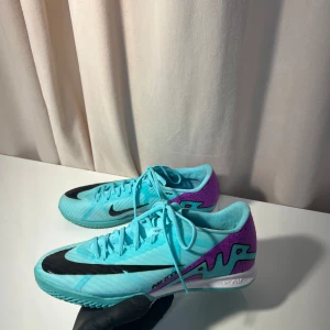 Nike fotbollsskor i turkos och lila - Snygga Nike fotbollsskor i en fräsch turkos färg med lila detaljer och svart Nike-logga. Skorna har snörning och en modern design som passar perfekt för fotbollsplanen.