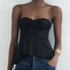 Svart korsettlinne med tunna axelband - Snygg svart korsettlinne med tunna axelband och volangdetalj nedtill. Toppen har en figurnära passform och är perfekt för en stilren look. Den har en elegant design med rynkade kupor och knytbara axelband. Aldrig använd helt ny med prislapp kvar💗nypris 399kr
