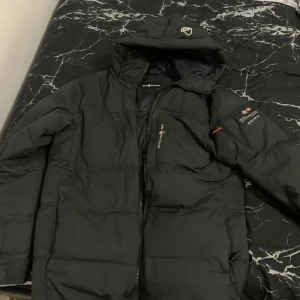 Svart dunjacka från Sail Racing - Säljer en svart dunjacka från Sail Racing med huva och dragkedja. Jackan har en stilren design med logotypdetaljer på ärmen och huvan. Perfekt för kalla dagar.