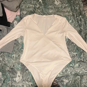 Vit långärmad bodysuit från H&M - Säljer en stilren vit bodysuit från H&M med lång ärm och v-ringning. Perfekt för att matcha med både jeans och kjol. Materialet är mjukt och stretchigt för en bekväm passform.
