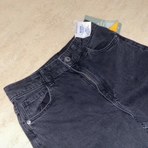 Svarta jeans - Oanvända jeans med hög midja och dragkedja. De har en klassisk design med fem fickor och knappstängning.