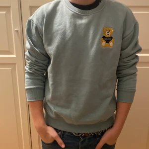 Grå sweatshirt från Moschino med nallemotiv - Säljer en stilren grå sweatshirt från Moschino med ett gulligt nallemotiv på bröstet. Tröjan har långa ärmar och ribbade muddar vid ärmslut och nederkant. Perfekt för en avslappnad look!