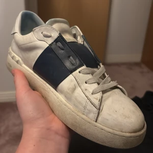 Valentino Garavani sneakers i vitt och blått - Snygga Valentino Garavani sneakers i vitt med en bred blå remsa över ovansidan. Skorna har snörning och en klassisk design som passar perfekt för en stilren look.