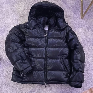Svart pufferjacka från Moncler - Säljer en snygg svart pufferjacka från Moncler med huva och dragkedja. Jackan har en quiltad design och långa ärmar. Perfekt för kyliga dagar.