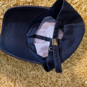 Mörkblå keps med justerbart spänne - Snygg mörkblå keps med justerbart spänne baktill för perfekt passform. Kepsen har en klassisk design och är tillverkad i ett slitstarkt material. Perfekt för både vardag och fritid.
