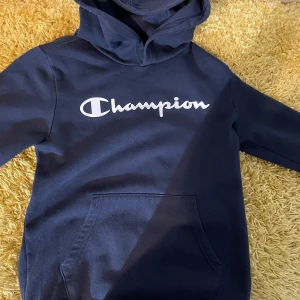 Mörkblå hoodie från Champion - Säljer en snygg mörkblå hoodie från Champion med deras ikoniska logga tryckt på bröstet. Tröjan har en klassisk känguruficka och en bekväm huva. Perfekt för en avslappnad stil.