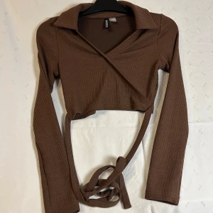 Brun ribbad omlottopp från Divided - Snygg brun ribbad omlottopp från Divided med långa ärmar och knytband i midjan. Perfekt för en stilren look. Passar bra till både jeans och kjol.