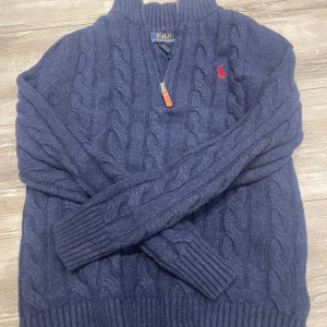 Blå stickad tröja från Ralph Lauren - Säljer en snygg blå stickad tröja från Ralph Lauren med kabelstickat mönster och en dragkedja vid halsen. Tröjan har långa ärmar och en liten röd logotyp på bröstet. Perfekt för kyliga dagar!