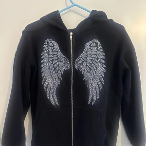 Svart hoodie med vingdetaljer från Shein - Säljer en svart hoodie från Shein med snygga vingdetaljer i silver på ryggen. Tröjan har dragkedja och långa ärmar, perfekt för en cool och avslappnad stil. Hör av dig vid intresse!